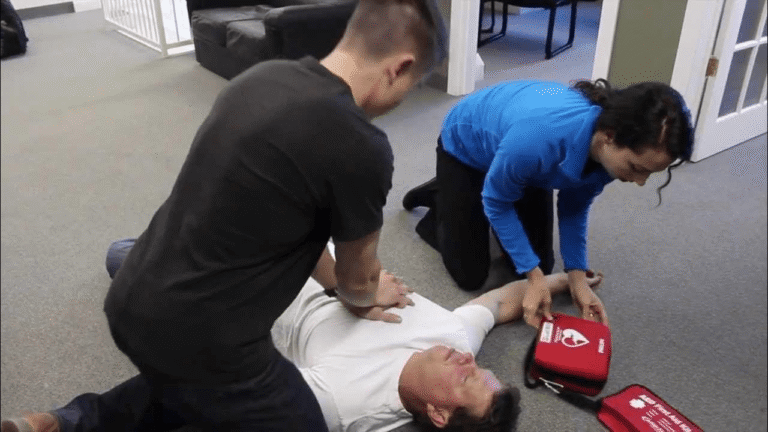 Two responders performing CPR and preparing an AED on an unconscious person during a first aid emergency.
